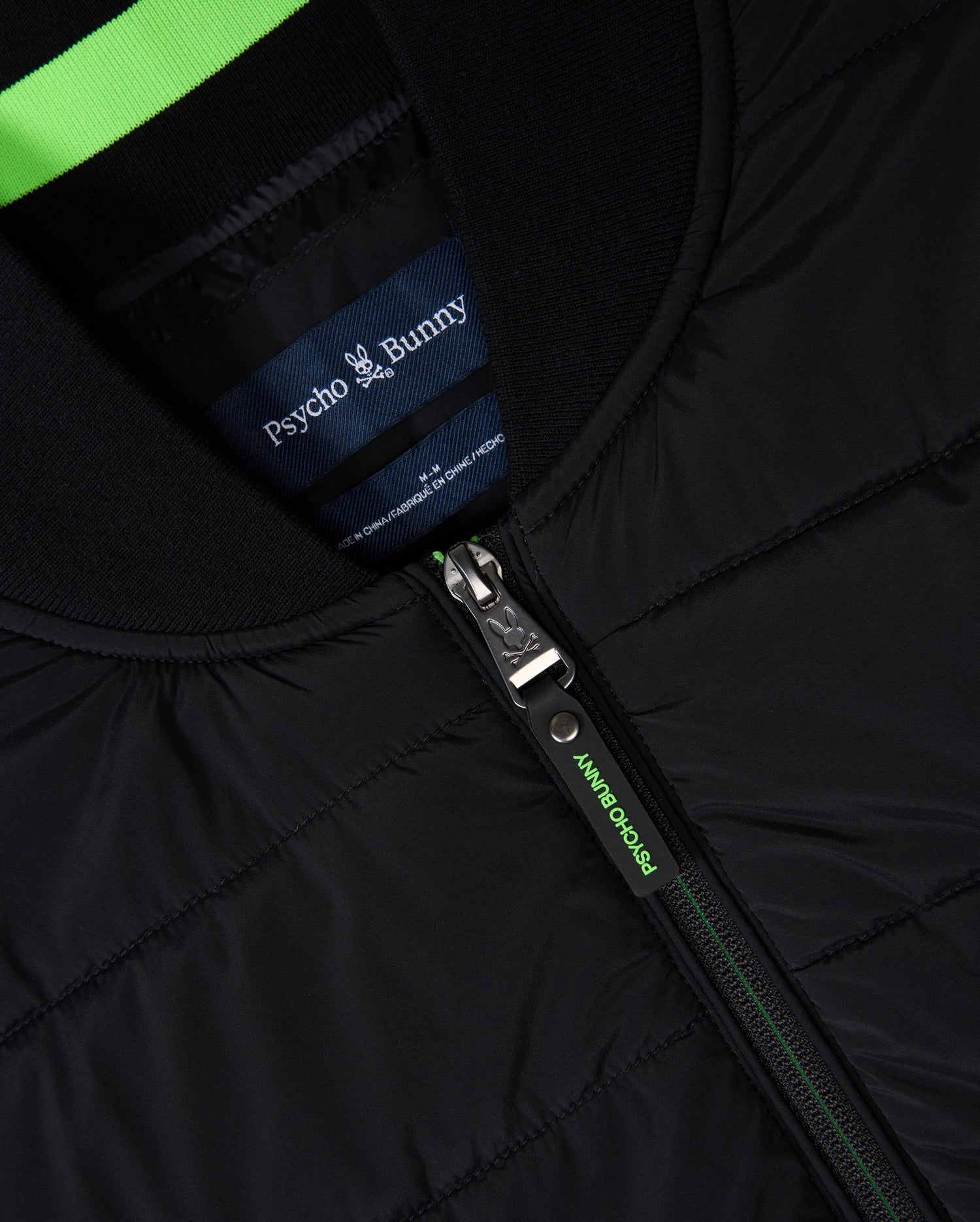 Close-up of a versatile MENS MYKEL MIXED MEDIA JACKET - B6J875D200 featuring a front zipper. The "Psycho Bunny" brand label adorns a blue and green striped tag inside the collar, while green accents along the zipper line add flair to this men's quilted design.