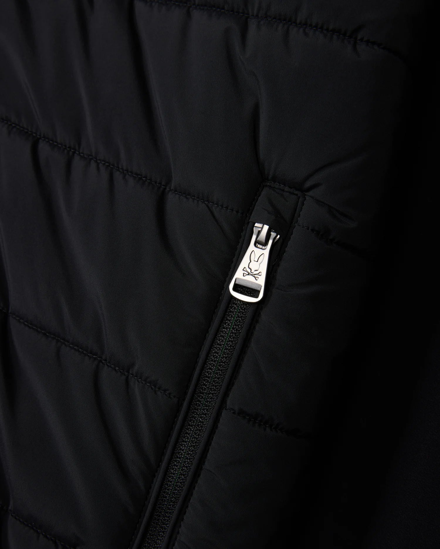 Close-up of the MENS MYKEL MIXED MEDIA JACKET by Psycho Bunny, showcasing a black quilted design with a silver zipper featuring an engraved logo. The versatile jacket includes horizontal line stitching for added texture.