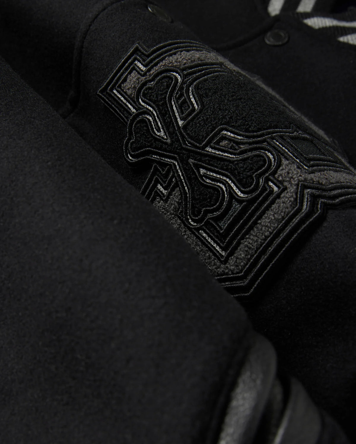 Close-up of the Psycho Bunny MENS BRAYDEN WOOL LEATHER VARSITY BOMBER - B6J876D200, showcasing an embroidered design with overlapping bones and intricate stitching. The thick, textured wool-blend fabric highlights the detailed craftsmanship of this black varsity jacket.