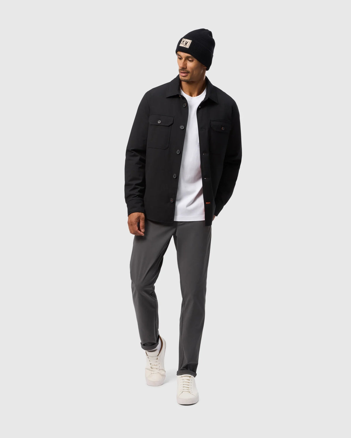 A man in a Psycho Bunny MENS HILLCREST PADDED SHACKET - B6J936D200 with patch pockets, a black beanie, white t-shirt, gray pants, and white sneakers stands against a light gray background. He looks to his left with one hand casually tucked in his pocket.