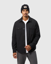 A man sports a Psycho Bunny MENS HILLCREST PADDED SHACKET (B6J936D200) with subtle neon lining over a white shirt and gray pants, topped off with a black beanie, against a plain gray background.