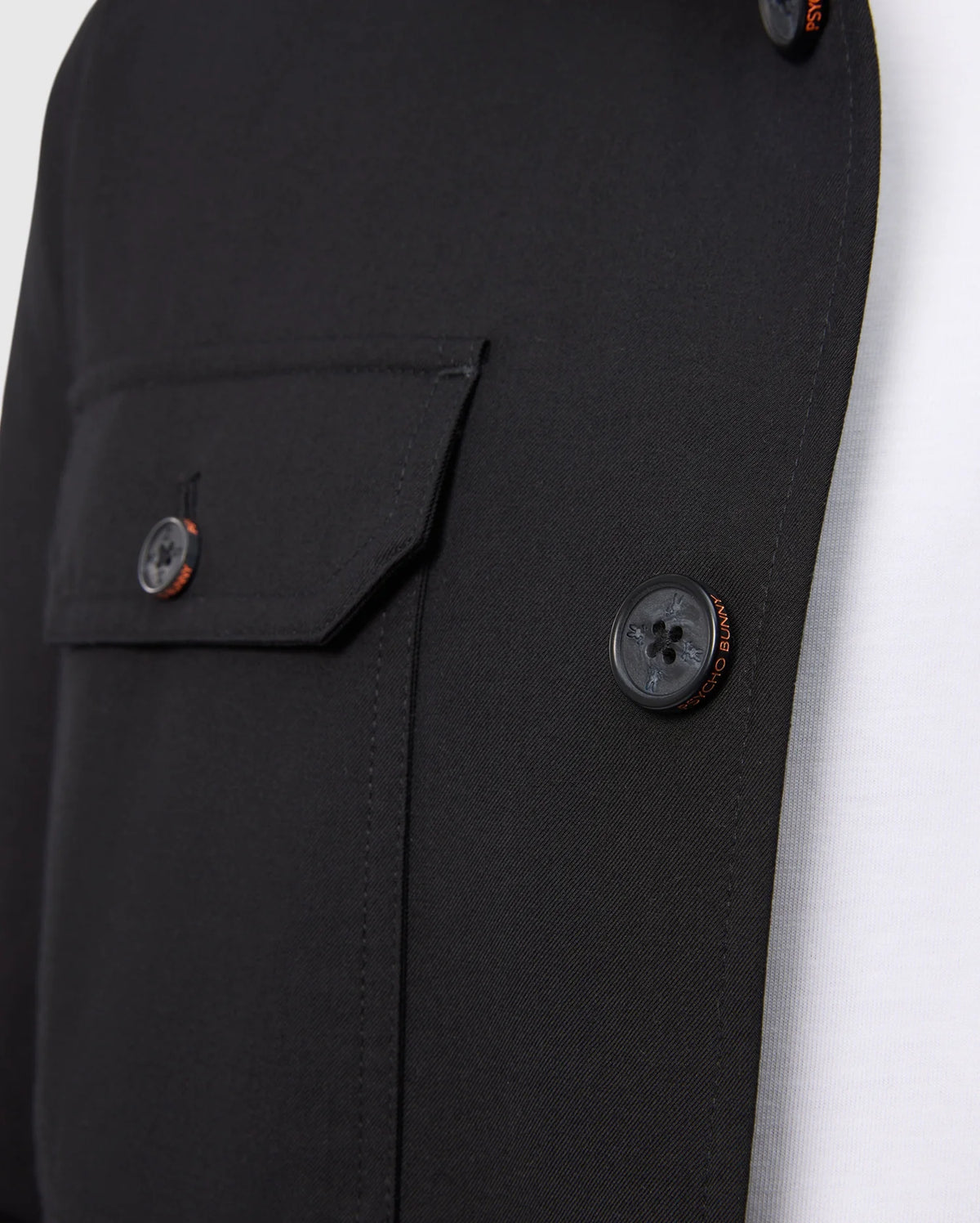 A close-up of the Psycho Bunny MENS HILLCREST PADDED SHACKET - B6J936D200, in black, showing visible buttons and a left flap pocket, layered over a sleek white shirt.