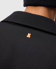 The image shows the back of a person wearing Psycho Bunny's Men's Hillcrest Padded Shacket, featuring a small orange bunny logo on the upper back. Their look is completed by a black knit hat, adding understated style to their ensemble.
