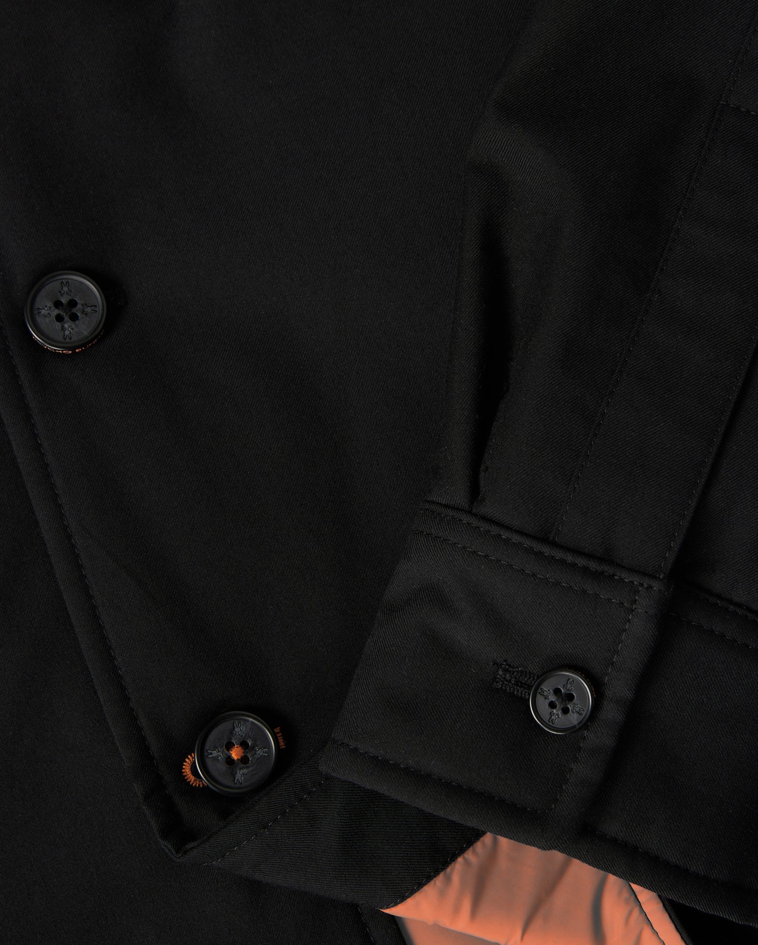 Close-up of the MENS HILLCREST PADDED SHACKET by Psycho Bunny, showcasing dark buttons, stitched buttonhole on the sleeve, patch pockets, and smooth black fabric with a hint of orange neon lining visible beneath the folded cuff.