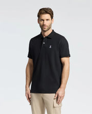A man with short hair and a beard wears the Psycho Bunny Men's Classic Pique Polo Shirt (B6K001ARPC) featuring diamond-knit piqué fabric and an embroidered logo. He stands against a plain light background in beige pants, looking neutral with his hands relaxed at his sides.