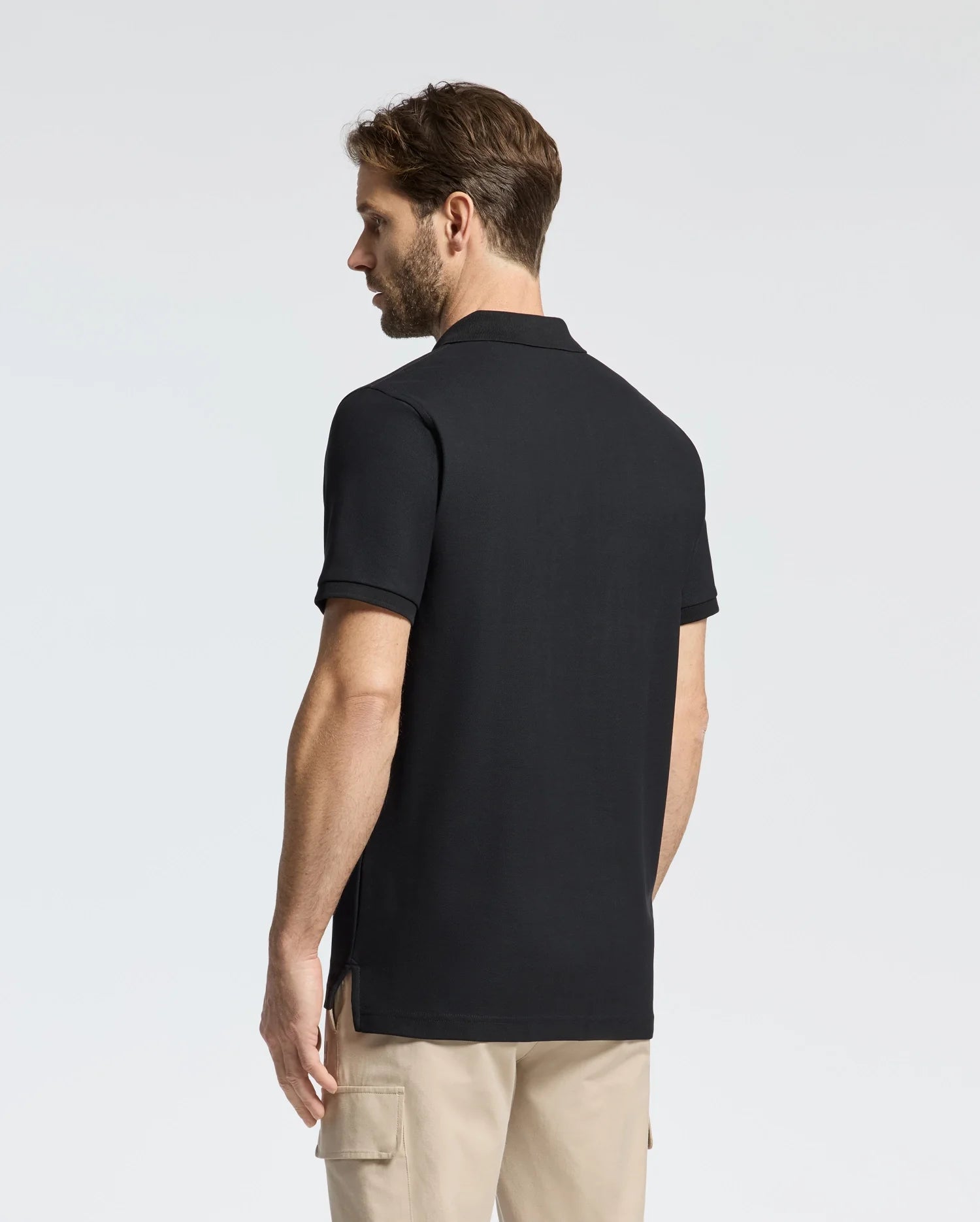 A man with short brown hair wears a Psycho Bunny men's classic black piqué polo shirt (B6K001ARPC) styled with light beige pants, standing and gazing left against a plain white background.