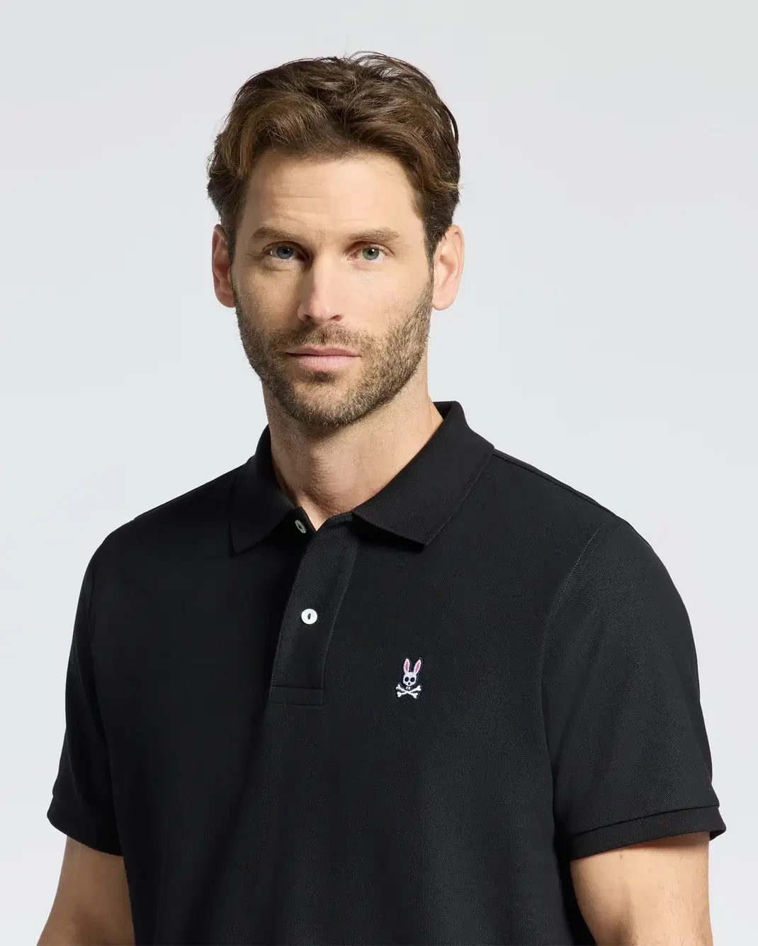 A man with short brown hair and a beard wears Psycho Bunny's black MENS CLASSIC PIQUE POLO SHIRT (B6K001ARPC) featuring a small embroidered bunny with crossbones. He has a neutral expression and stands against a plain, light gray background.