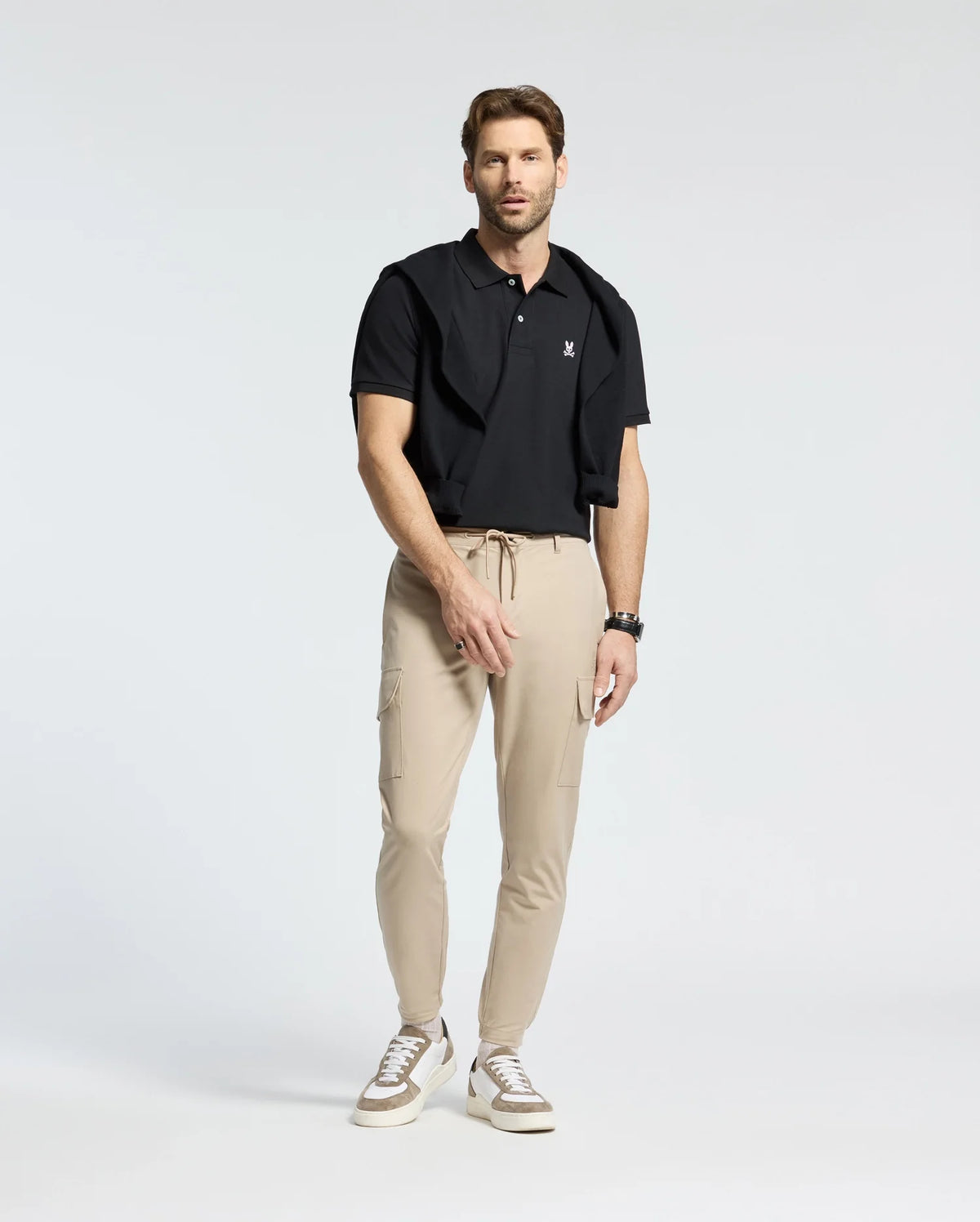 A brown-haired man stands in a studio wearing Psycho Bunny's Men's Classic Pique Polo Shirt in black, with a black sweater draped over his shoulders, beige cargo pants, and white sneakers. He wears a watch on his left wrist and looks forward against a plain white background.