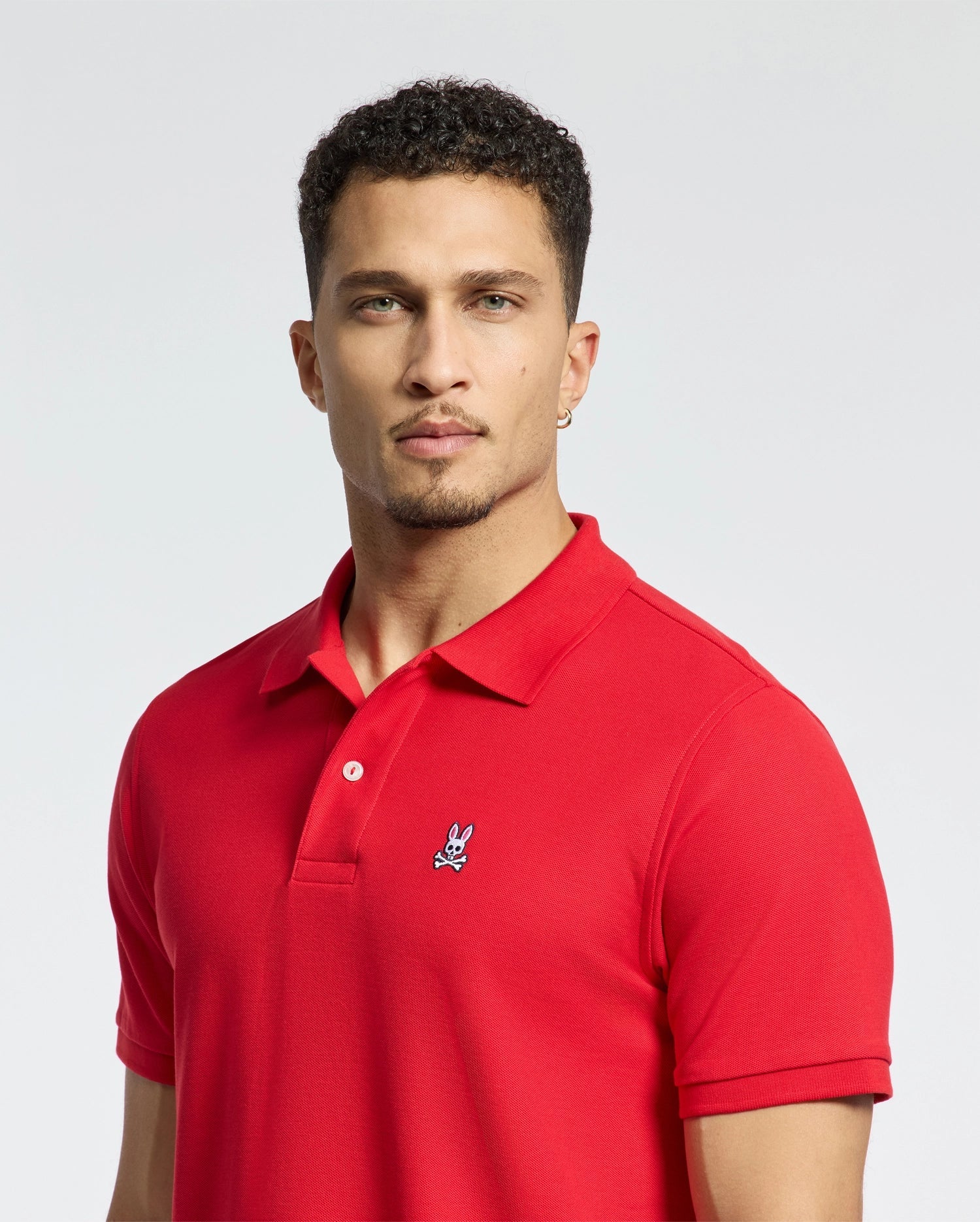 A man with short curly hair wears a vibrant red Psycho Bunny Men's Classic Pique Polo Shirt, crafted from soft Pima cotton and adorned with mother-of-pearl buttons and a small embroidered logo. The plain, light-colored background accentuates the vibrancy of his clothing.