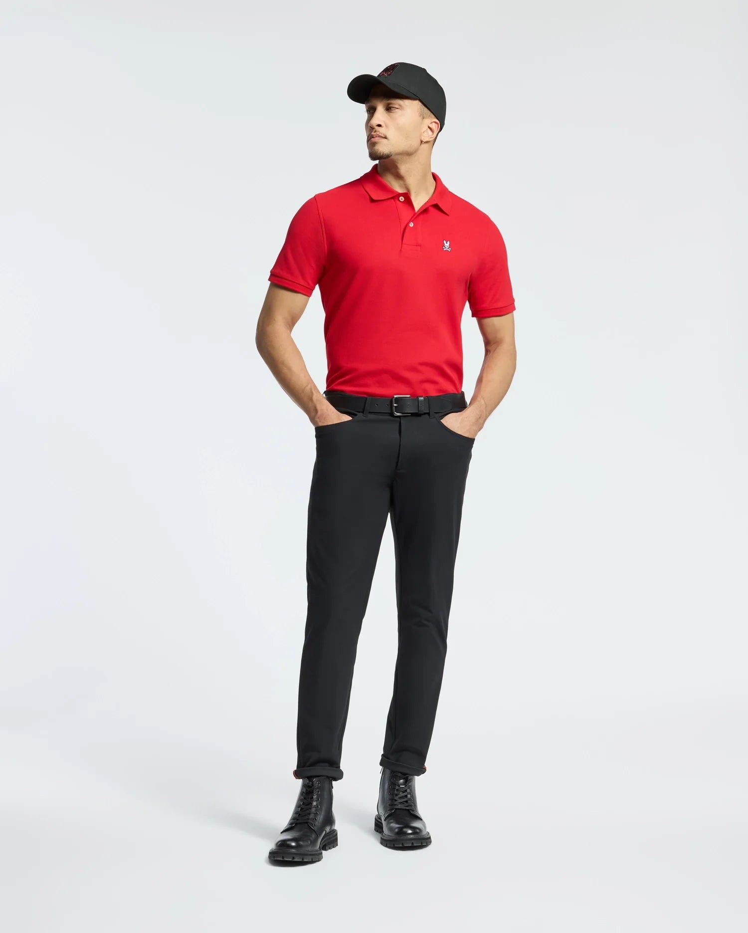 A person confidently stands with hands in pockets against a plain white background, wearing a Psycho Bunny Men's Classic Pique Polo Shirt (B6K001CRPC) made of Pima cotton, paired with black pants and black boots. The bold red shirt is highlighted by mother-of-pearl buttons, adding an elegant touch to the outfit.