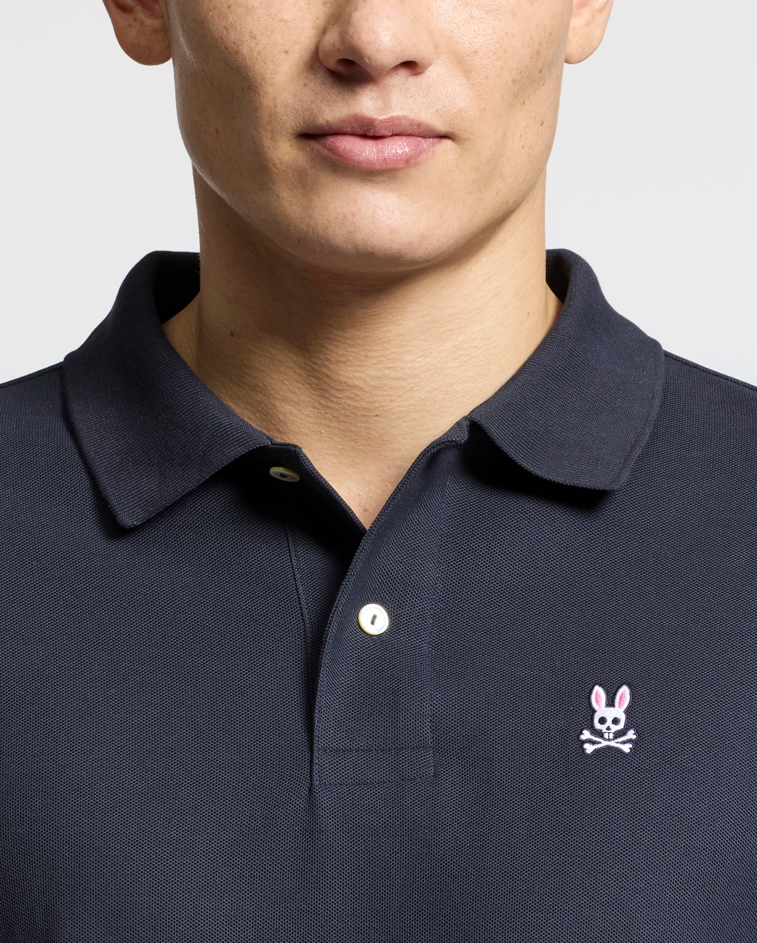 A man wears the Psycho Bunny Men's Classic Pique Polo Shirt - B6K001ARPC, made from soft Pima cotton in a dark color, featuring a pink-eared bunny skull emblem on crossed bones. The buttoned collar stands out against the plain white background.