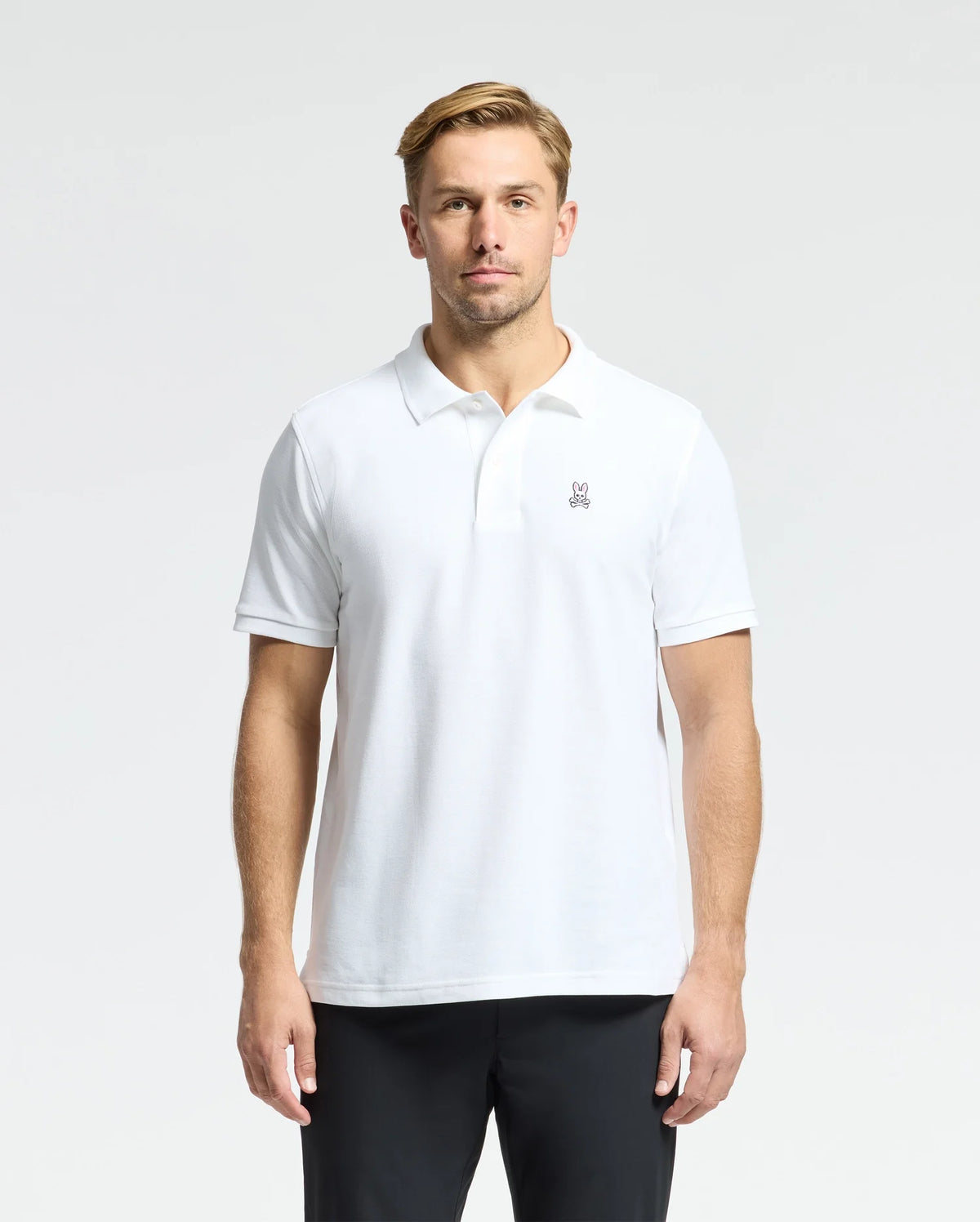 A man in a MENS CLASSIC PIQUE POLO SHIRT by Psycho Bunny, made from 100% Pima cotton and featuring mother-of-pearl buttons, stands against a plain background. His hands rest casually at his sides, and he wears a neutral expression.