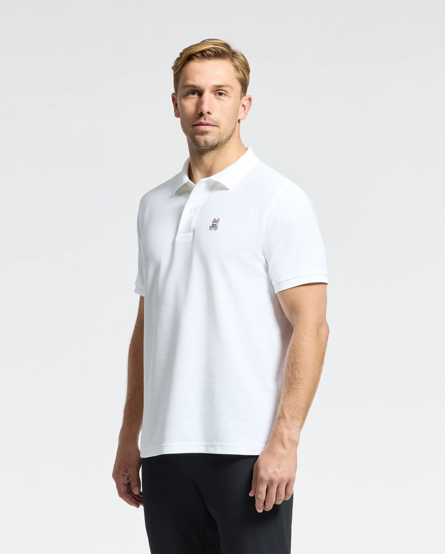 A person dressed in a Psycho Bunny MENS CLASSIC PIQUE POLO SHIRT - B6K001ARPC, crafted from 100% Pima cotton and featuring mother-of-pearl buttons along with a small embroidered logo on the chest, stands against a plain background. They have short blond hair and are looking at the camera.