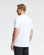 A man with short blond hair is standing with his back to the camera. He is dressed in a Psycho Bunny MENS CLASSIC PIQUE POLO SHIRT - B6K001ARPC, featuring mother-of-pearl buttons, paired with black pants, all set against a plain white background.