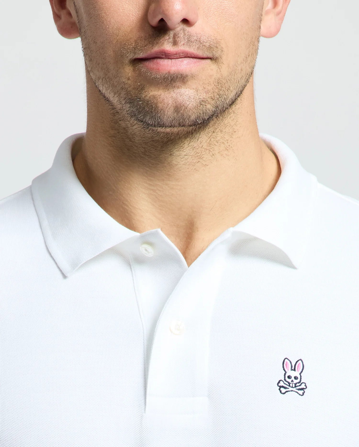 A man is wearing a Psycho Bunny Men's Classic Pique Polo Shirt - B6K001ARPC, made from 100% Pima cotton. It features a subtle embroidered bunny and crossbones logo on the chest and elegant mother-of-pearl buttons. The plain gray backdrop enhances the shirt's sophisticated simplicity.