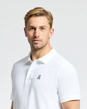 A man with short blond hair is wearing the Psycho Bunny MENS CLASSIC PIQUE POLO SHIRT - B6K001ARPC, made from 100% Pima cotton and adorned with a small embroidered rabbit logo on the chest. He gazes directly at the camera against a light grey background.