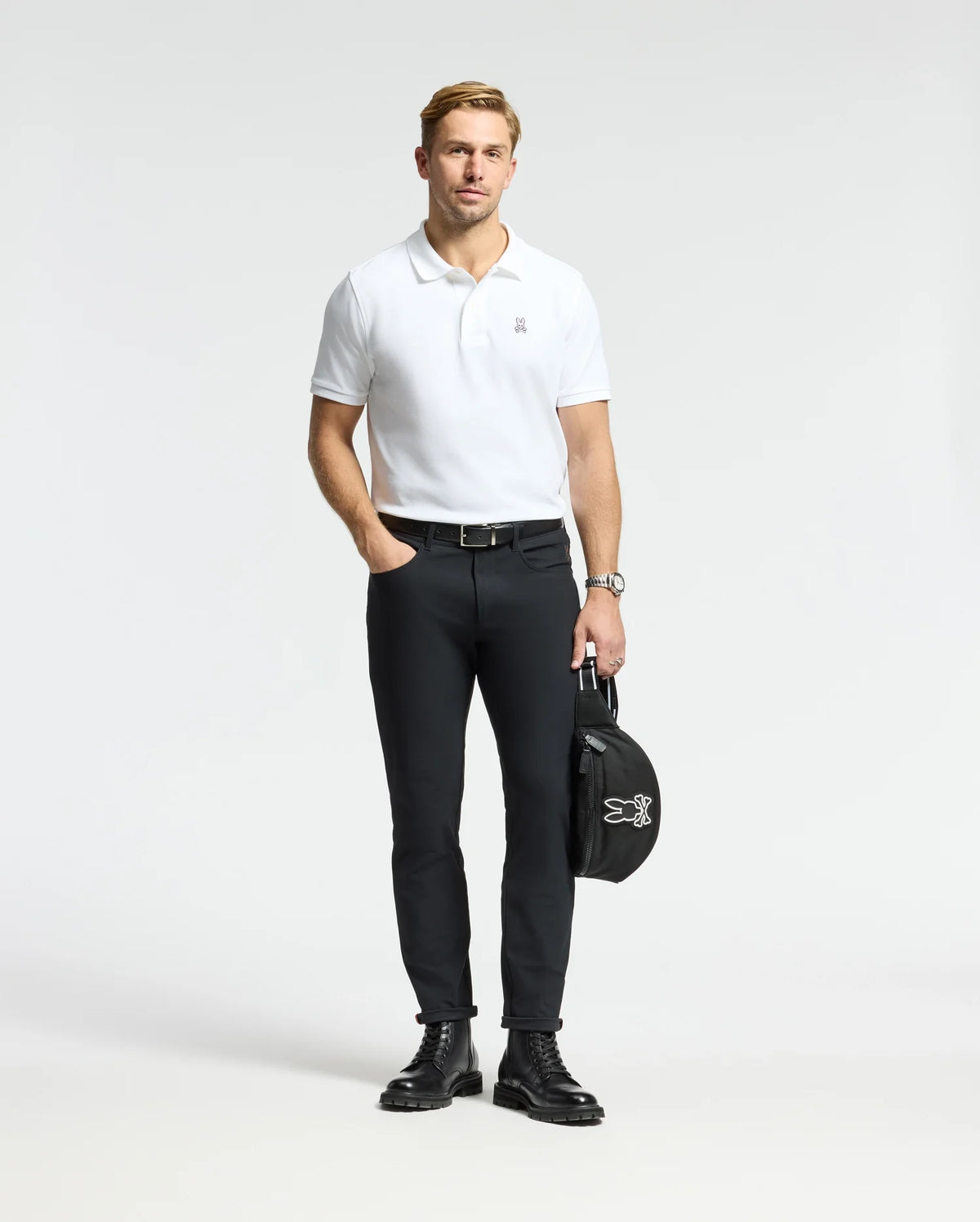 A person stands wearing a Psycho Bunny MENS CLASSIC PIQUE POLO SHIRT - B6K001ARPC, crafted from 100% Pima cotton, paired with black pants. They hold a black jacket featuring a white logo in their left hand against a plain, light-colored background. The ensemble is completed with black boots and a watch on their left wrist that accentuates the mother-of-pearl buttons.