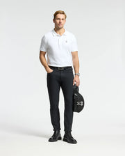 A person stands wearing a Psycho Bunny MENS CLASSIC PIQUE POLO SHIRT - B6K001ARPC, crafted from 100% Pima cotton, paired with black pants. They hold a black jacket featuring a white logo in their left hand against a plain, light-colored background. The ensemble is completed with black boots and a watch on their left wrist that accentuates the mother-of-pearl buttons.