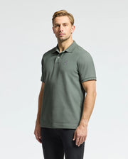 A person clad in the MENS CLASSIC PIQUE POLO SHIRT - B6K001B200 by Psycho Bunny, adorned in green with an embroidered Bunny logo on the chest, stands against a plain light background. They sport short hair and are paired with black pants made from soft Pima cotton.