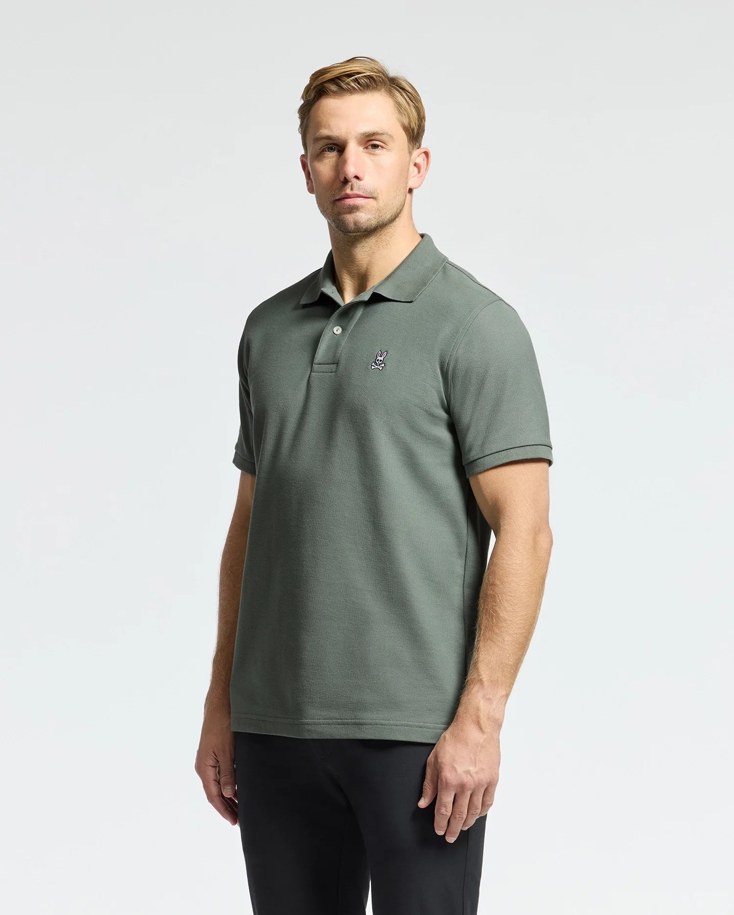 A person clad in the MENS CLASSIC PIQUE POLO SHIRT - B6K001B200 by Psycho Bunny, adorned in green with an embroidered Bunny logo on the chest, stands against a plain light background. They sport short hair and are paired with black pants made from soft Pima cotton.
