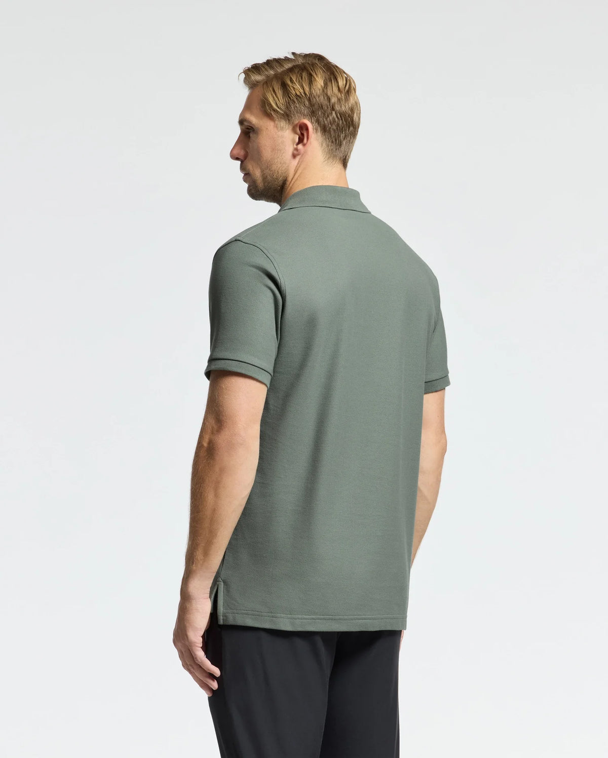 A man with blonde hair is wearing a green Psycho Bunny MENS CLASSIC PIQUE POLO SHIRT - B6K001B200, which features the embroidered Bunny logo, and black pants. He stands with his back to the camera against a plain white background.