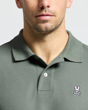 A person wears the Psycho Bunny MENS CLASSIC PIQUE POLO SHIRT - B6K001B200 in olive green, showcasing an embroidered Bunny logo with crossed bones on the left chest. Made from premium Pima cotton, the plain background highlights the distinctive design of this unique apparel.
