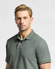 A man with short, light brown hair is wearing the Psycho Bunny MENS CLASSIC PIQUE POLO SHIRT - B6K001B200 in green, which features an embroidered bunny logo. He stands against a neutral background, looking directly at the camera with a neutral expression.