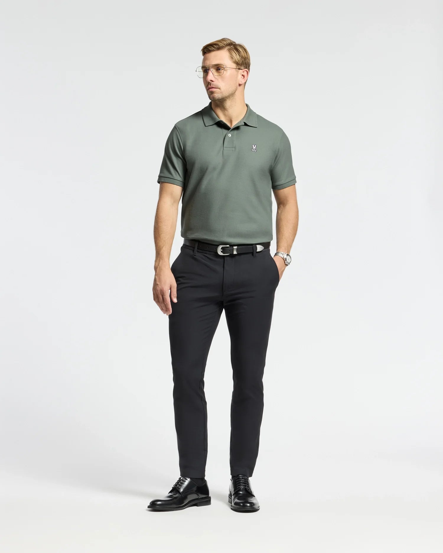 A man stands wearing the Psycho Bunny MENS CLASSIC PIQUE POLO SHIRT - B6K001B200, crafted from Pima cotton and showcasing the embroidered Bunny logo. He pairs it with black pants and shoes. With short hair, glasses, and a watch on his left wrist, he poses against a plain white background.