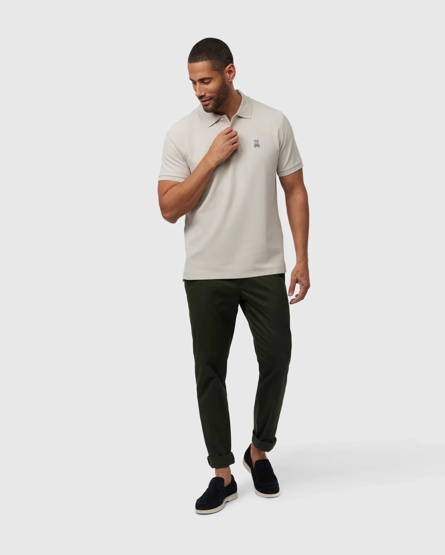 A man wearing a timeless men's Psycho Bunny MENS CLASSIC PIQUE POLO SHIRT - B6K001B200 in light grey, dark green pants, and black loafers stands against a plain white background. He looks down and to his right while adjusting the collar of his luxurious Pima cotton polo with his right hand.