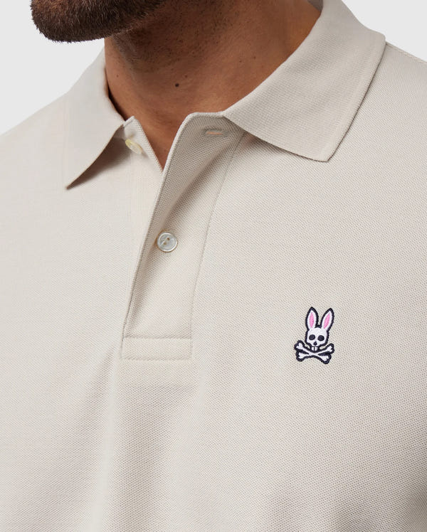 Close-up shot of a person wearing a light beige Psycho Bunny MENS CLASSIC PIQUE POLO SHIRT - B6K001B200 with a collar and buttons. This timeless shirt, crafted from luxurious Pima cotton, features an embroidered logo on the left chest area depicting a pink bunny head above crossed bones. Only part of the person’s face and neck are visible.