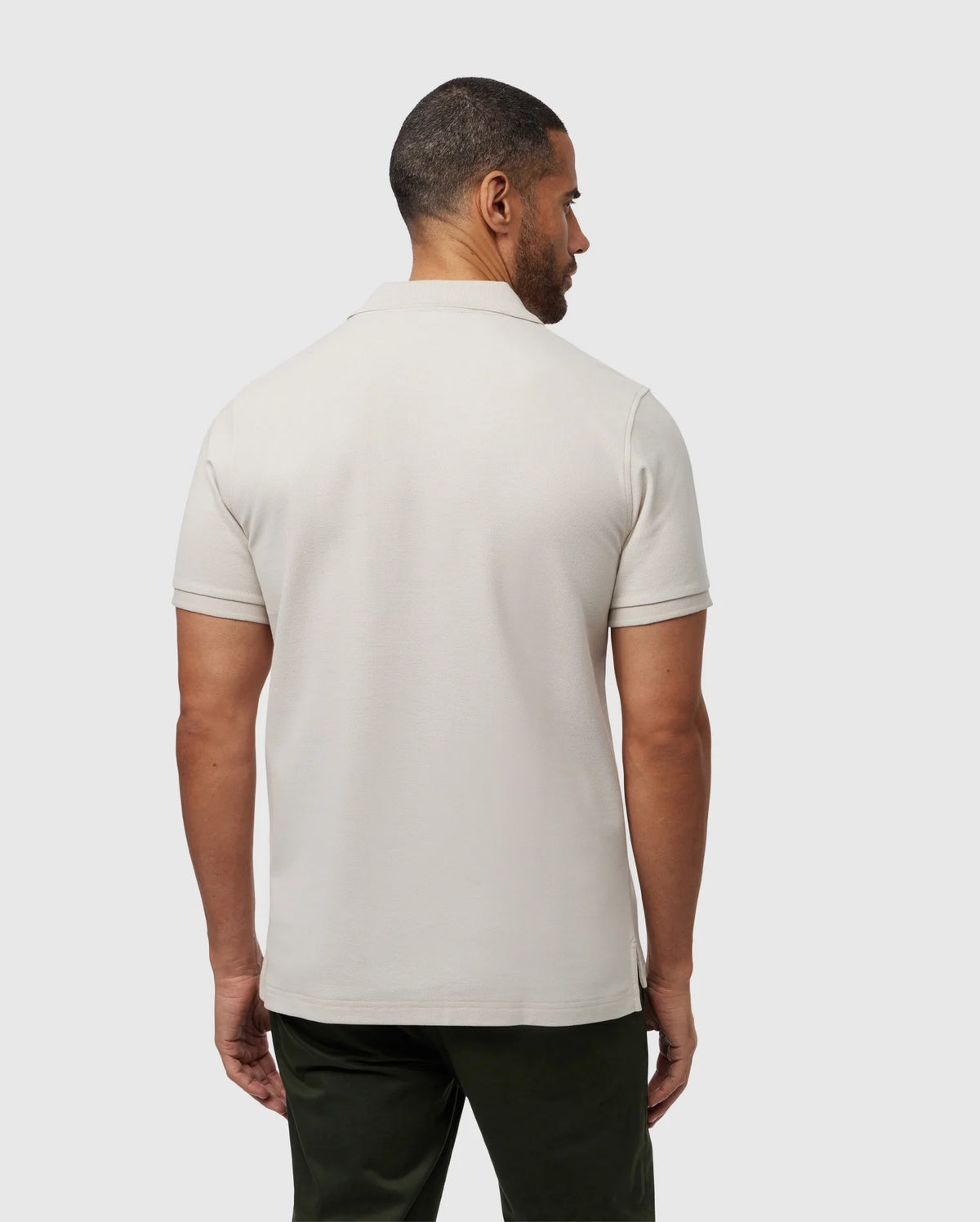 A man with short hair is standing with his back to the camera, showcasing the perfect fit of his light beige MENS CLASSIC PIQUE POLO SHIRT - B6K001B200 by Psycho Bunny. He pairs it effortlessly with dark green pants against a plain, light gray background.