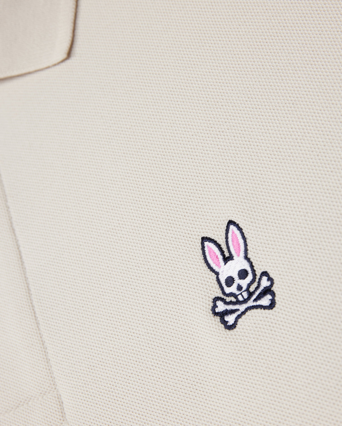Close-up of a beige textured fabric from the Psycho Bunny MENS CLASSIC PIQUE POLO SHIRT - B6K001B200, with an embroidered logo featuring a white skull and crossbones with pink-eared bunny ears on the chest area. Crafted from luxurious Pima cotton for a perfect fit.