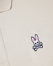 Close-up of a beige textured fabric from the Psycho Bunny MENS CLASSIC PIQUE POLO SHIRT - B6K001B200, with an embroidered logo featuring a white skull and crossbones with pink-eared bunny ears on the chest area. Crafted from luxurious Pima cotton for a perfect fit.