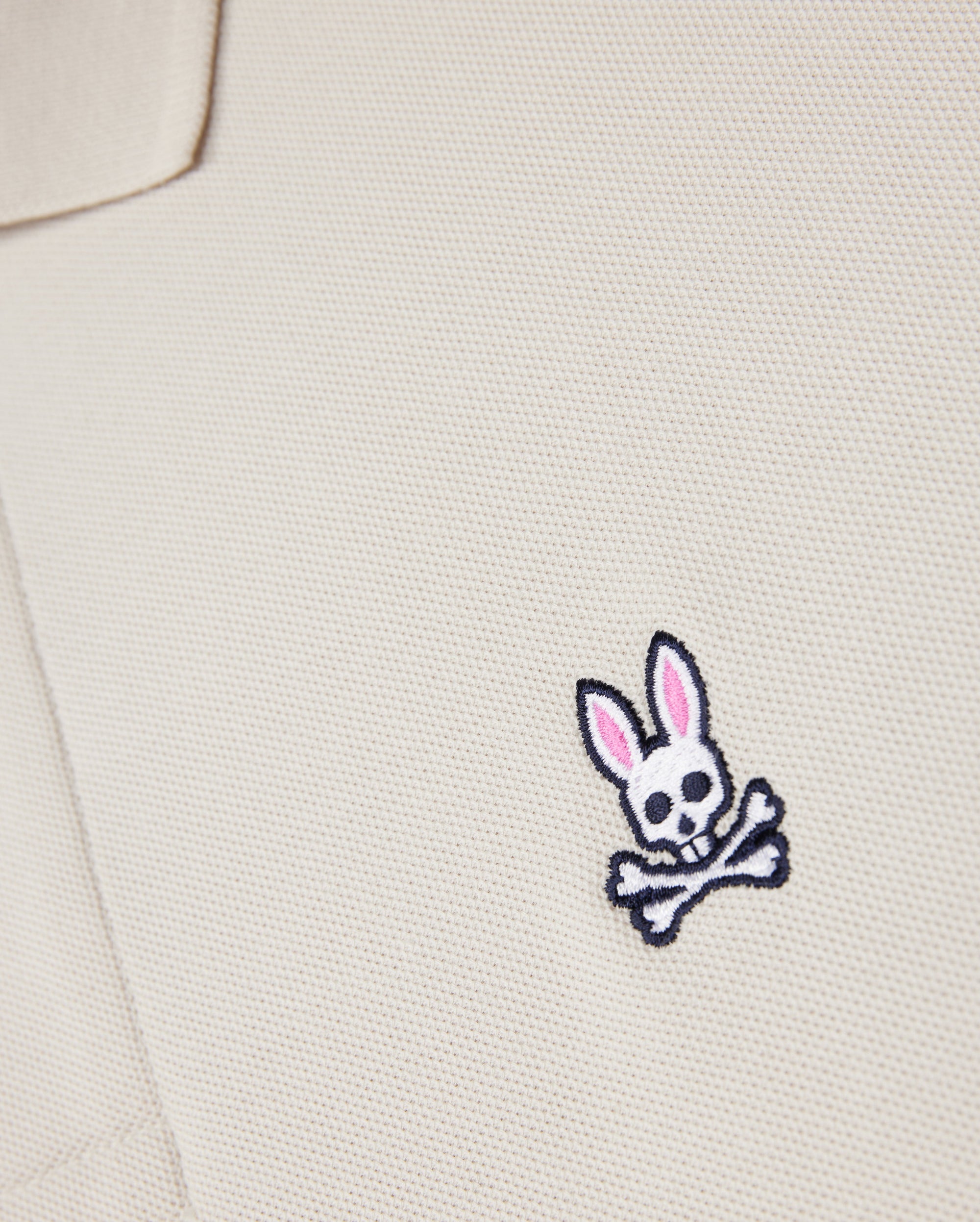 Close-up of a beige textured fabric from the Psycho Bunny MENS CLASSIC PIQUE POLO SHIRT - B6K001B200, with an embroidered logo featuring a white skull and crossbones with pink-eared bunny ears on the chest area. Crafted from luxurious Pima cotton for a perfect fit.