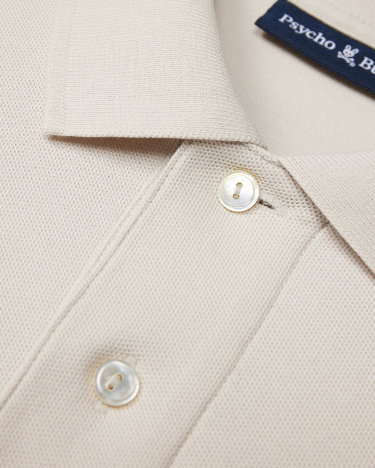 Close-up of a beige Psycho Bunny MENS CLASSIC PIQUE POLO SHIRT - B6K001B200 collar with two white buttons. The timeless shirt fabric has a textured, waffle-like pattern. A navy blue label with white text is partially visible near the collar, hinting at its luxurious Pima cotton composition.