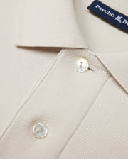 Close-up of a beige Psycho Bunny MENS CLASSIC PIQUE POLO SHIRT - B6K001B200 collar with two white buttons. The timeless shirt fabric has a textured, waffle-like pattern. A navy blue label with white text is partially visible near the collar, hinting at its luxurious Pima cotton composition.