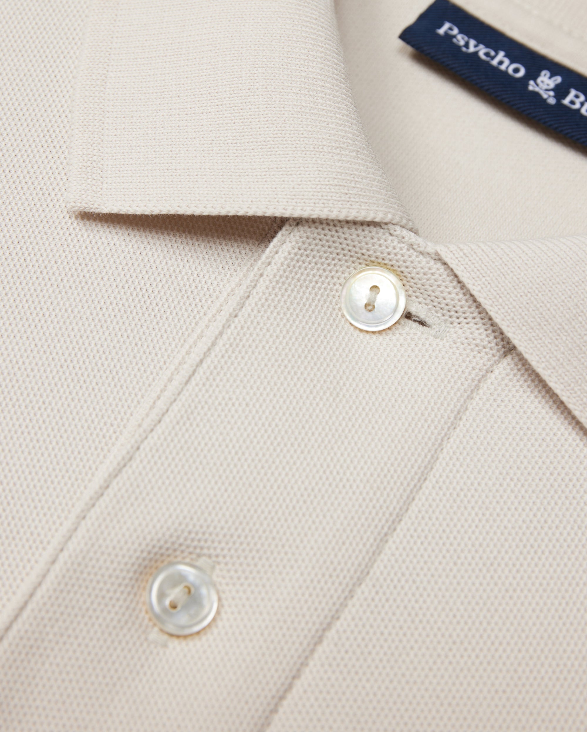 Close-up of a beige Psycho Bunny MENS CLASSIC PIQUE POLO SHIRT - B6K001B200 collar with two white buttons. The timeless shirt fabric has a textured, waffle-like pattern. A navy blue label with white text is partially visible near the collar, hinting at its luxurious Pima cotton composition.