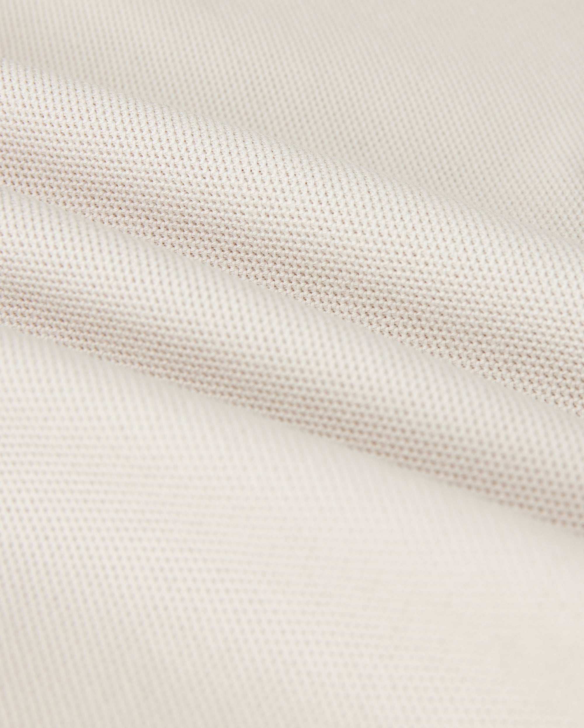 Close-up of a softly folded, light beige textured fabric with a subtle grid pattern, showcasing its smooth and pliable nature. The gentle folds add depth and highlight the luxurious Pima cotton's fine weave and soft sheen, hinting at the perfect fit for a Psycho Bunny Men's Classic Pique Polo Shirt - B6K001B200.