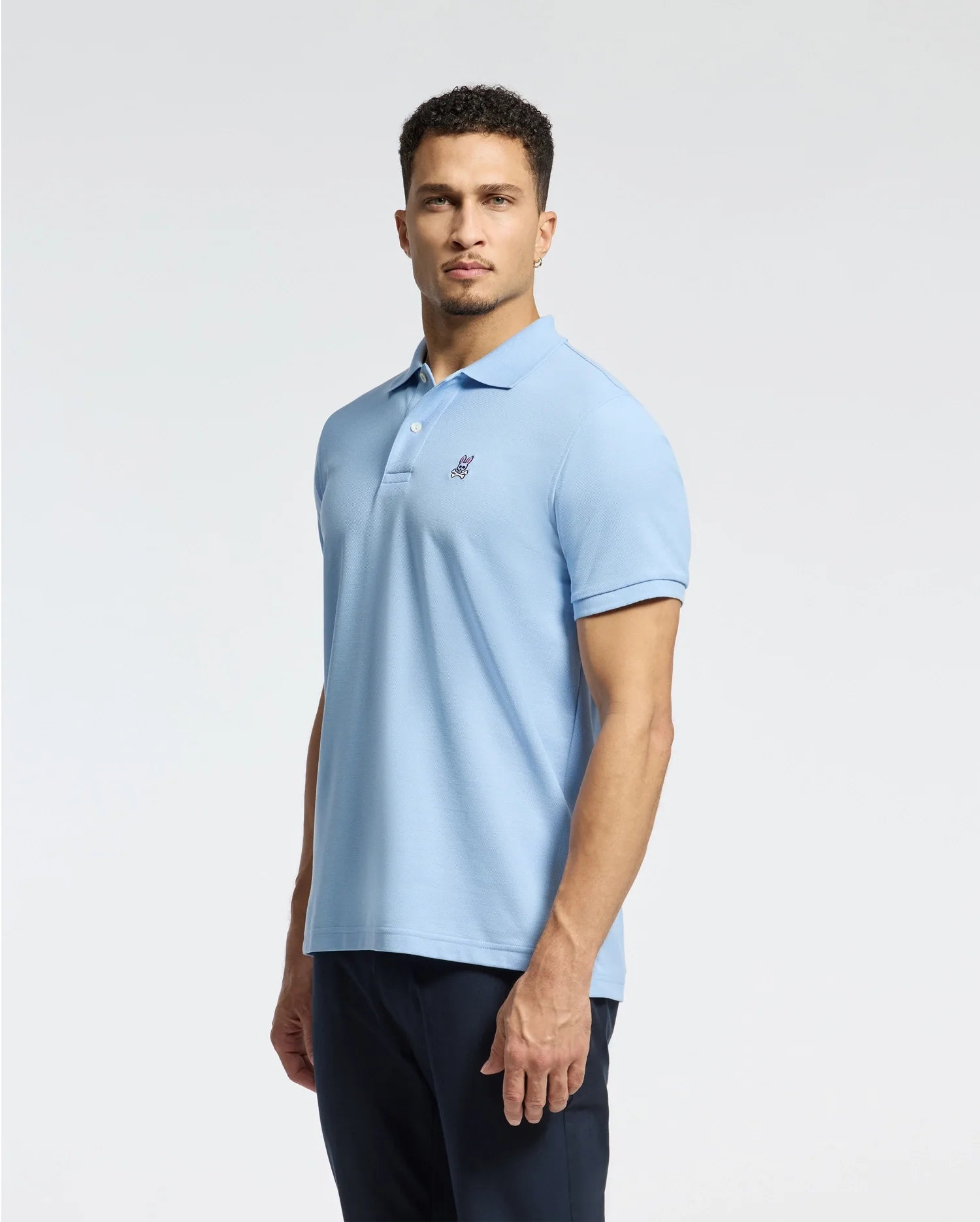 A man stands wearing a MENS CLASSIC PIQUE POLO SHIRT - B6K001B200 from Psycho Bunny in light blue and dark pants against a plain white background. The polo, crafted from soft Pima cotton, is adorned with a small embroidered Bunny logo on the chest.