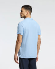 A man with short curly hair faces away from the camera, dressed in a light blue Psycho Bunny MENS CLASSIC PIQUE POLO SHIRT - B6K001B200, which features an embroidered Bunny logo. He pairs it with dark pants against a plain, light-colored background.