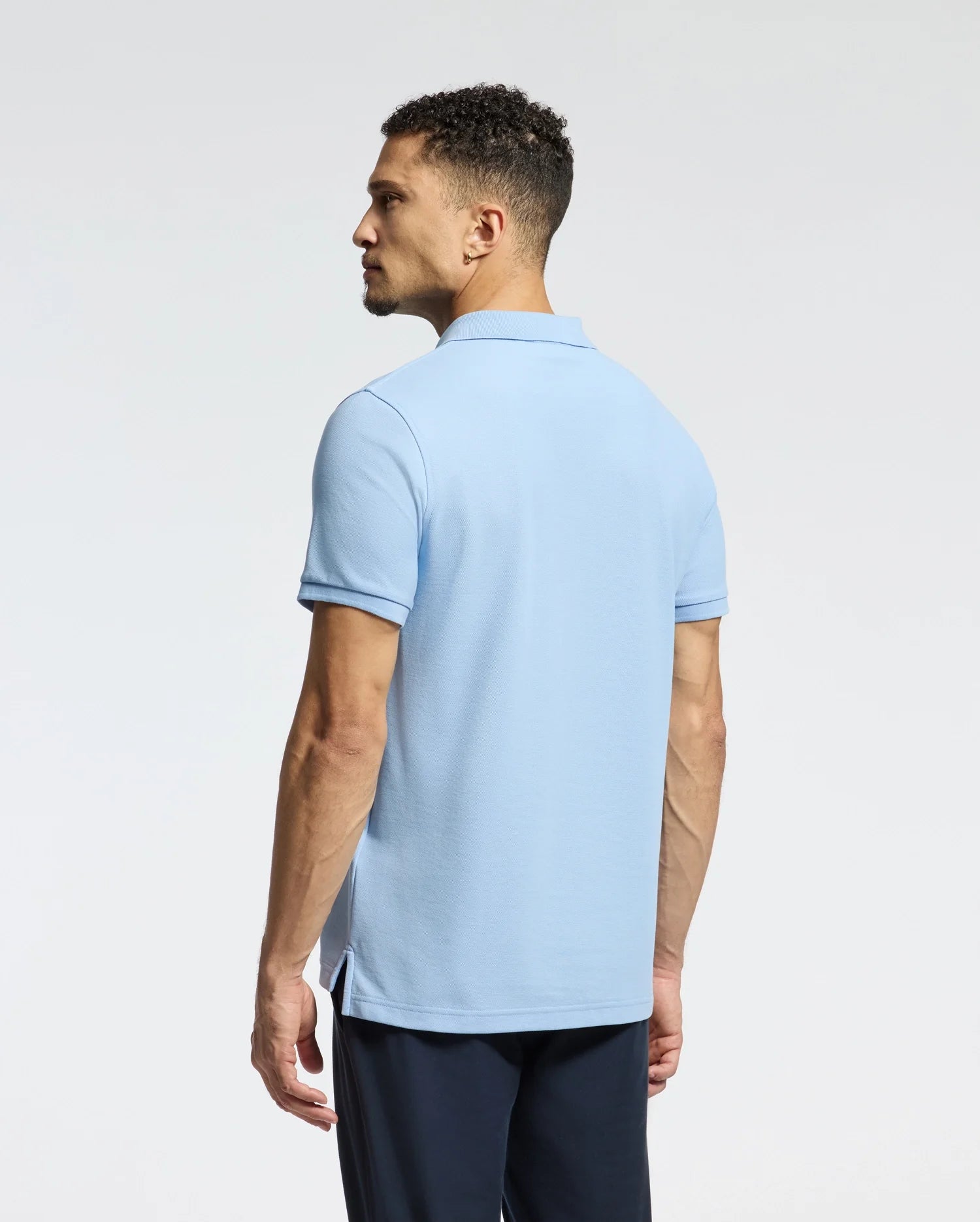 A man with short curly hair faces away from the camera, dressed in a light blue Psycho Bunny MENS CLASSIC PIQUE POLO SHIRT - B6K001B200, which features an embroidered Bunny logo. He pairs it with dark pants against a plain, light-colored background.