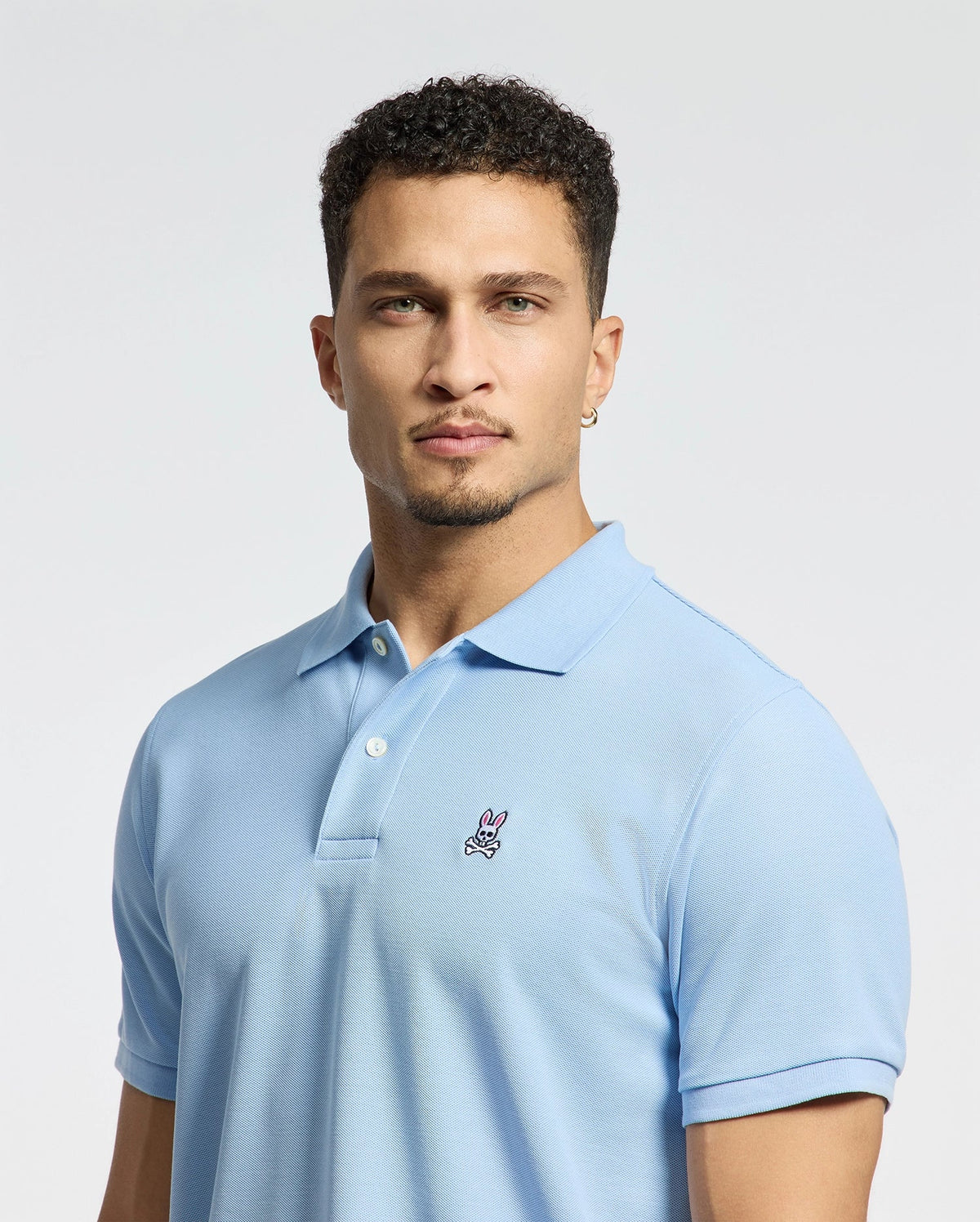 A person with short curly hair and a light beard is wearing the Psycho Bunny Men's Classic Piqué Polo Shirt in light blue, featuring an embroidered Bunny logo on the chest. The background is plain white.