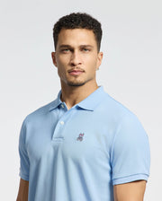 A person with short curly hair and a light beard is wearing the Psycho Bunny Men's Classic Piqué Polo Shirt in light blue, featuring an embroidered Bunny logo on the chest. The background is plain white.