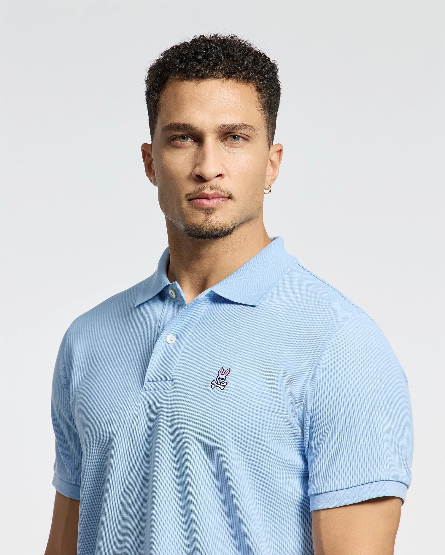 A person with short curly hair and a light beard is wearing the Psycho Bunny Men's Classic Piqué Polo Shirt in light blue, featuring an embroidered Bunny logo on the chest. The background is plain white.