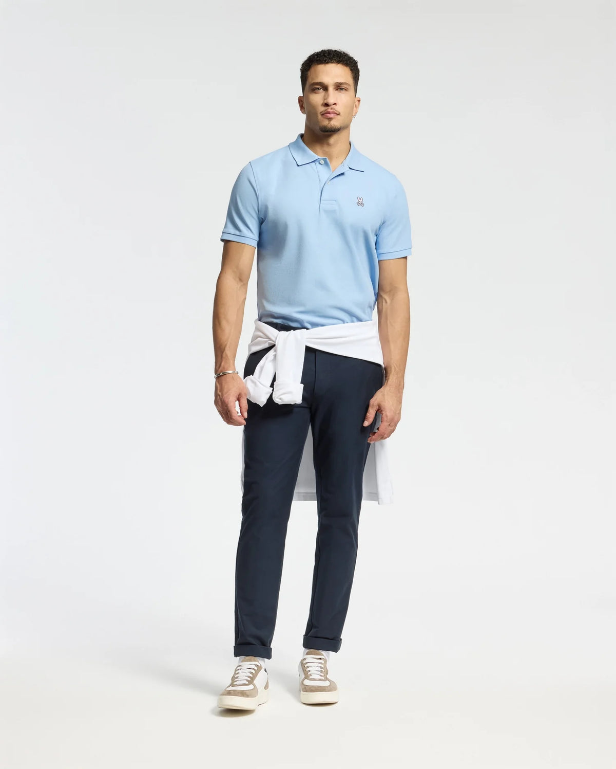 A man exudes confidence, donning a Psycho Bunny MENS CLASSIC PIQUE POLO SHIRT crafted from soft Pima cotton, which he pairs with dark pants and a white sweater casually tied around his waist. Light brown sneakers add the finishing touch to his ensemble, while the subtle embroidered Bunny logo enhances the charm of his attire against a simple white backdrop.