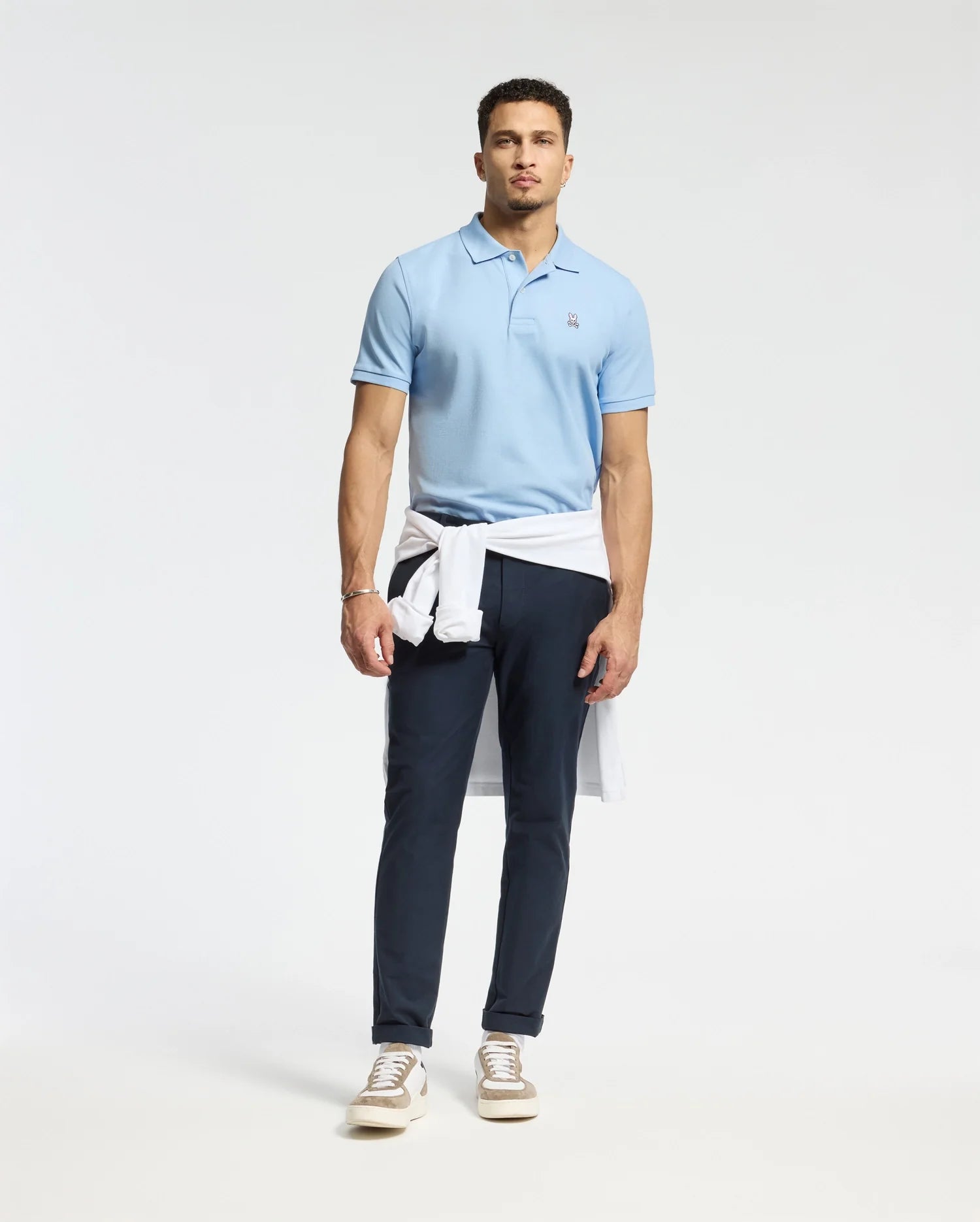 A man exudes confidence, donning a Psycho Bunny MENS CLASSIC PIQUE POLO SHIRT crafted from soft Pima cotton, which he pairs with dark pants and a white sweater casually tied around his waist. Light brown sneakers add the finishing touch to his ensemble, while the subtle embroidered Bunny logo enhances the charm of his attire against a simple white backdrop.