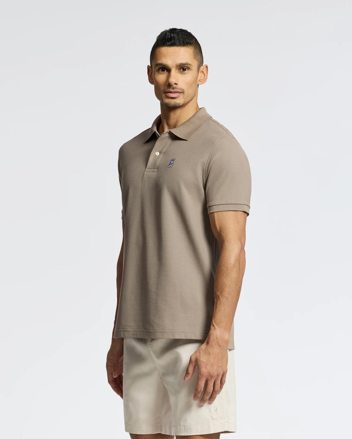 A man with short dark hair wears a taupe Psycho Bunny MENS CLASSIC PIQUE POLO SHIRT - B6K001CRPC, featuring a subtle embroidered logo, and beige shorts, standing against a plain white background.