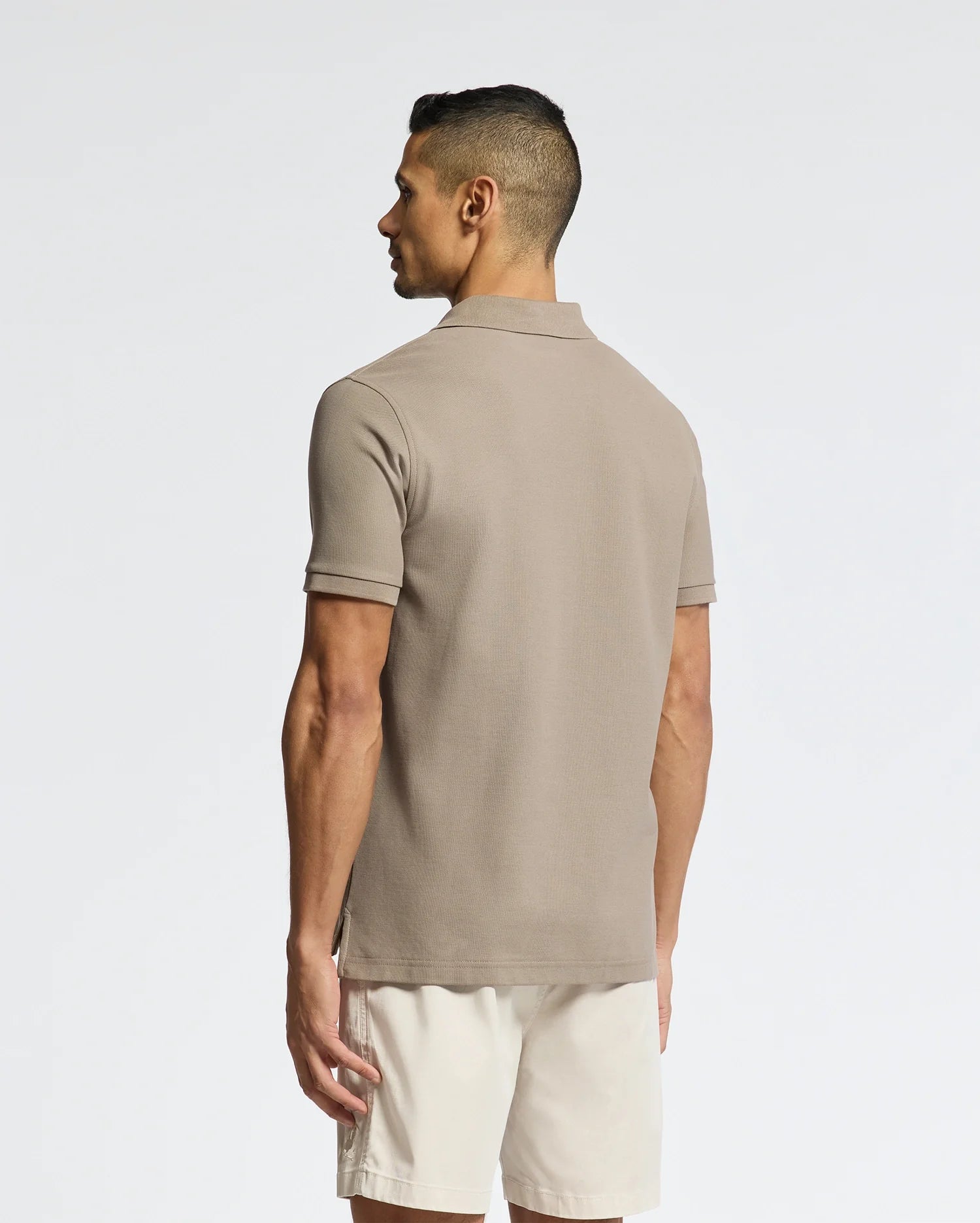 A man with short dark hair, facing away, wears a Psycho Bunny MEN'S CLASSIC PIQUE POLO SHIRT (B6K001CRPC) in taupe paired with light-colored shorts against a plain white background.