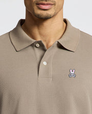 A man in a Psycho Bunny Men's Classic Pique Polo Shirt (B6K001CRPC) in beige displays the brand's small crossbones logo on the left chest, with his face partly visible.