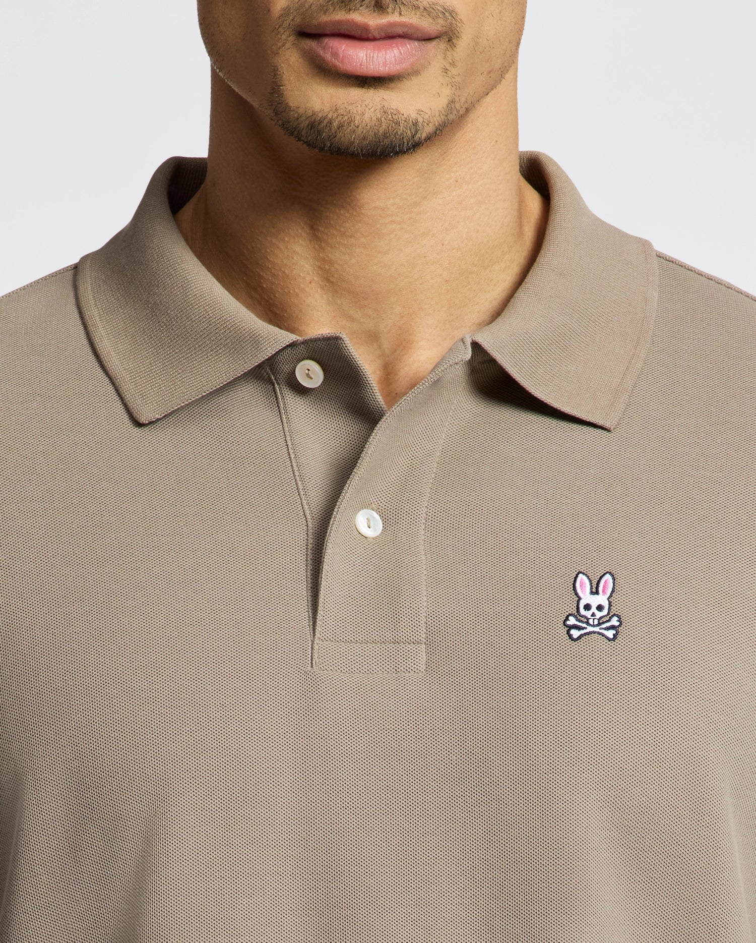 A man in a Psycho Bunny Men's Classic Pique Polo Shirt (B6K001CRPC) in beige displays the brand's small crossbones logo on the left chest, with his face partly visible.